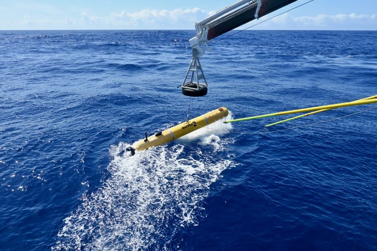 A yellow torpedo shaped autonomous underwater vehicle is dropped into a blue ocean with a launch and recovery system. 
