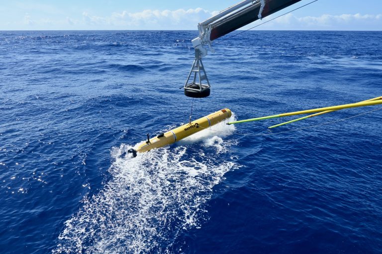 A yellow torpedo shaped autonomous underwater vehicle is dropped into a blue ocean with a launch and recovery system.