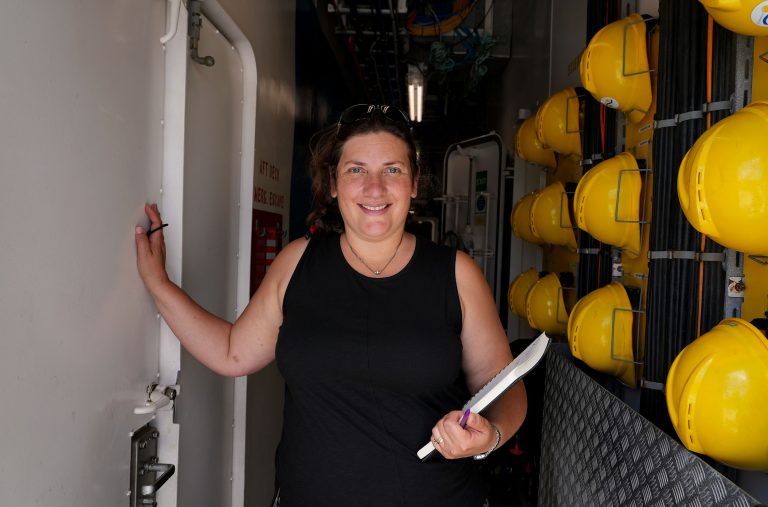 A picture of the Chief Scientist, Randi Rotjan in a hallway. She is holding a notebook. To her left are yellow hardhats.