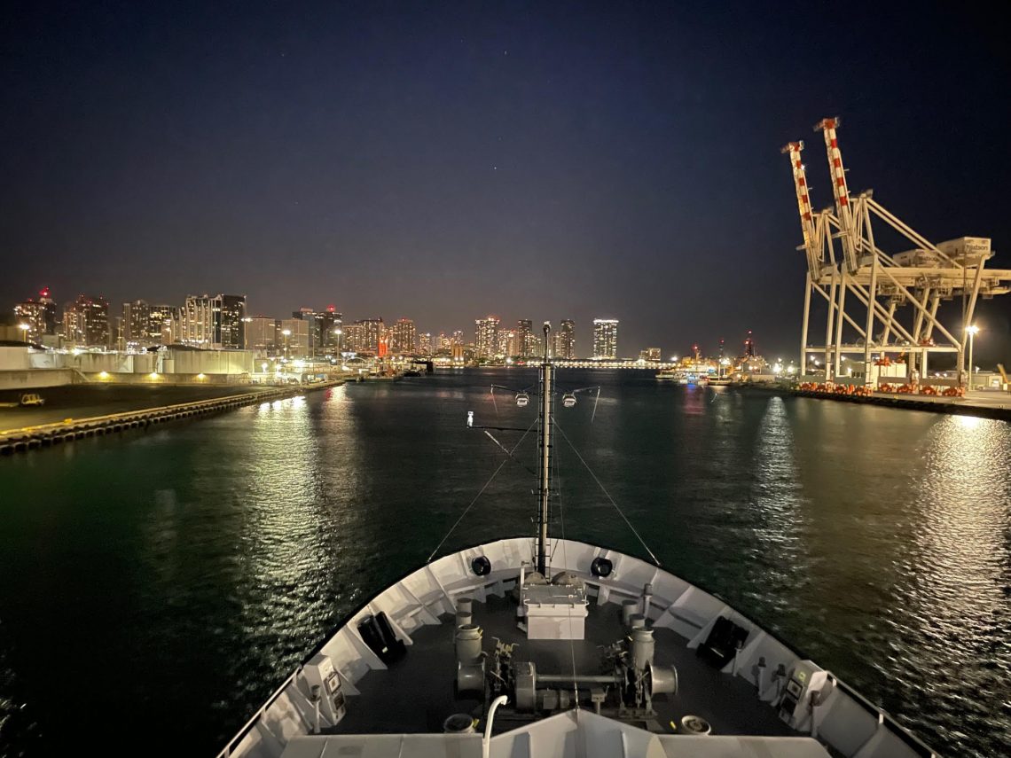 The bow of R/V Falkor surrounded by the Honolulu Harbor. The picture was taken late at night so the water and sky are dark and the city is lit up.