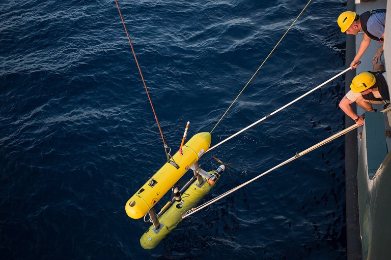 Crew prepare to retrieve AUV Sirius after a mission. This is accomplished by using poles to attach lines to it via poles then lifting the vehicle on deck via crane.